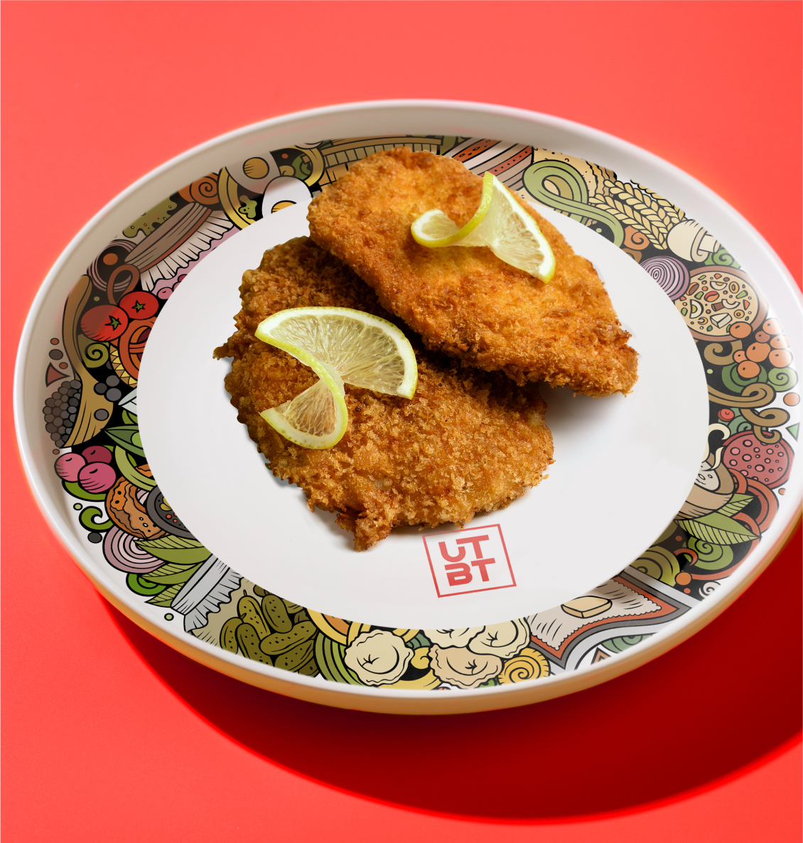Golden fried cutlets with lime wedges on UTBT branded plate with food illustrations against red background