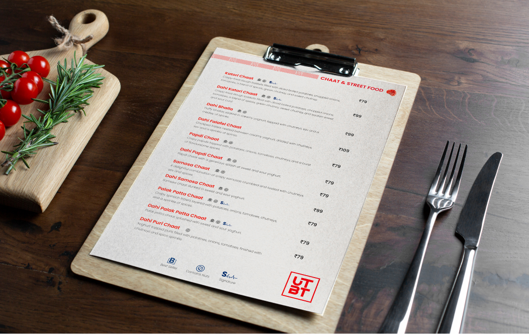 UTBT restaurant chaat menu on clipboard with utensils, cutting board, tomatoes and herbs on wooden table