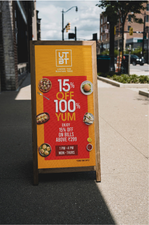 UTBT street advertising board showing 15% off and 100% YUM promotion on urban sidewalk with buildings background