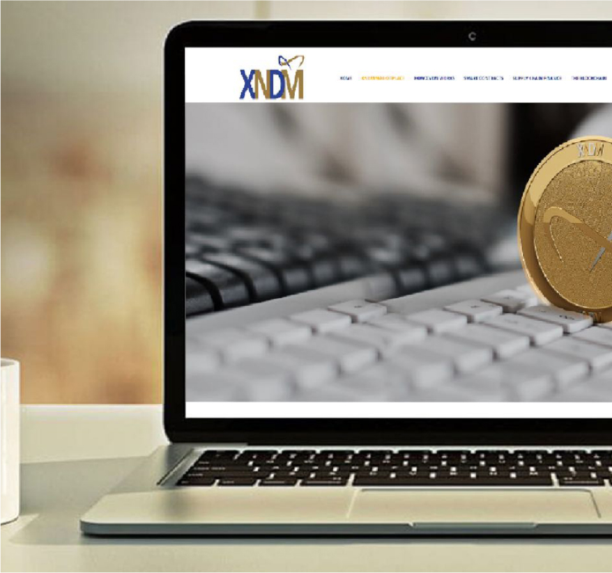 Laptop displaying XNDM website with gold cryptocurrency coin on keyboard, showing blockchain marketplace interface.
