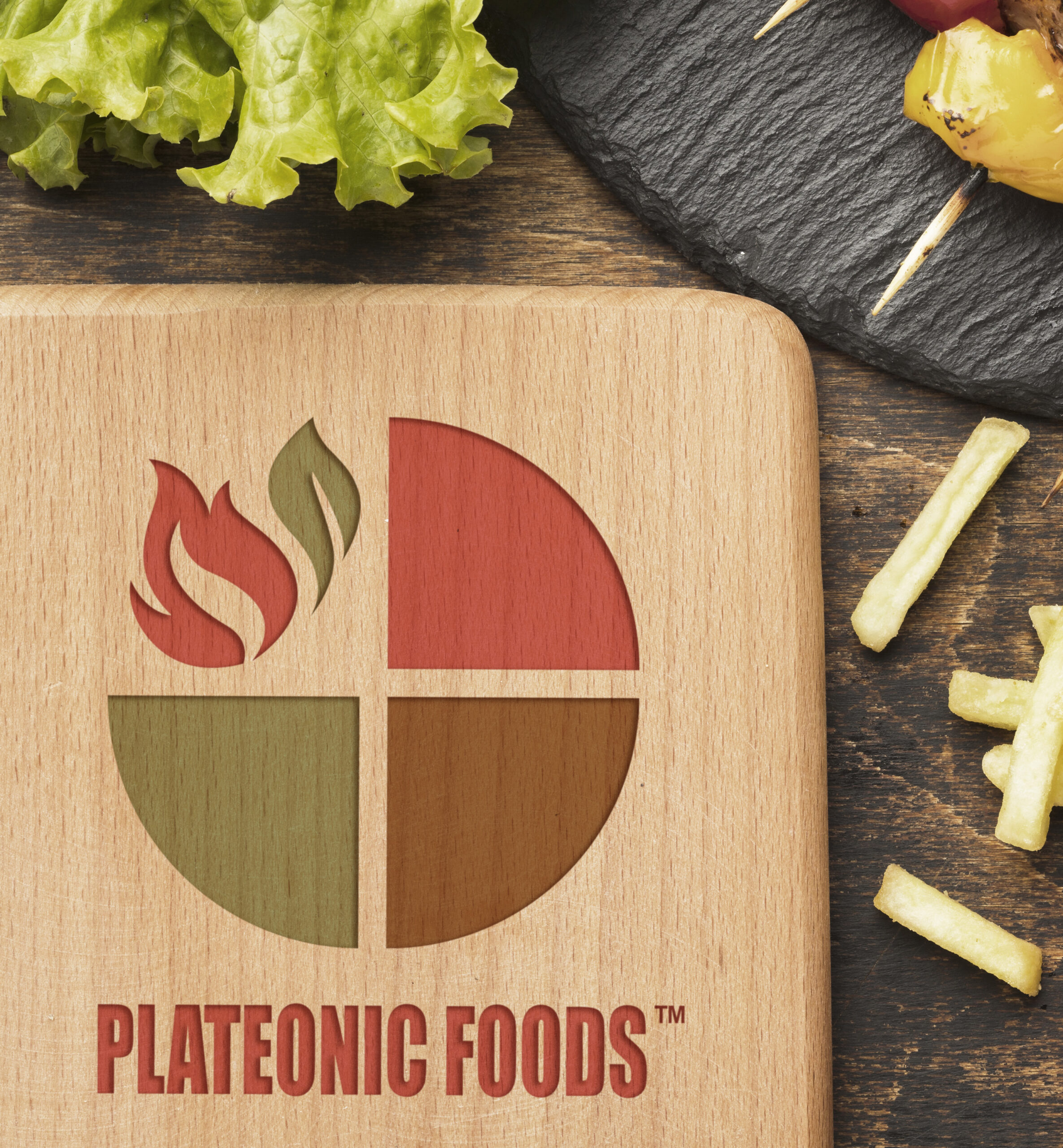 Plateonic Foods logo engraved on a wooden board, with lettuce, fries, and grilled vegetables in the background.