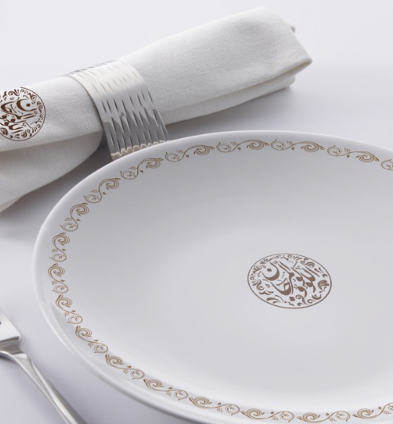 Elegant white plate with gold Arabic calligraphy and scroll border, paired with a napkin and silver napkin ring.