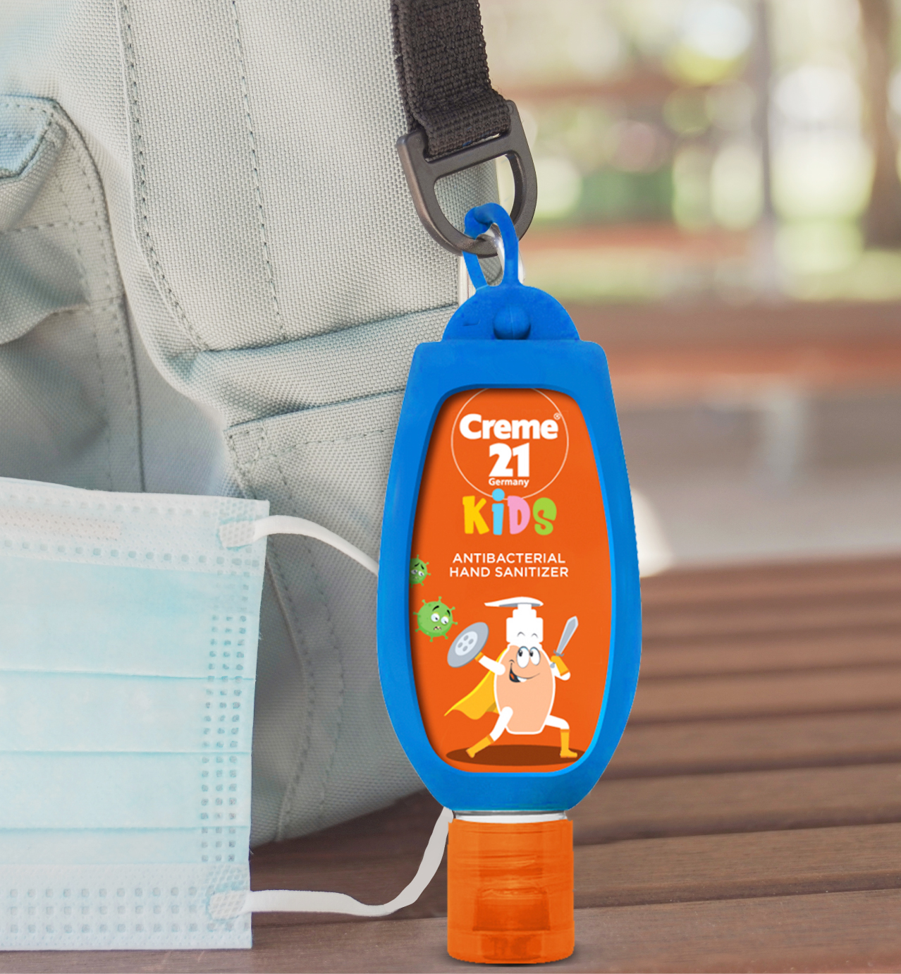 Creme 21 Kids antibacterial hand sanitizer in a blue holder clipped to a backpack, next to a disposable face mask.