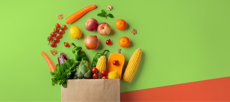 Paper grocery bag overflowing with fresh fruits and vegetables on green and orange background.