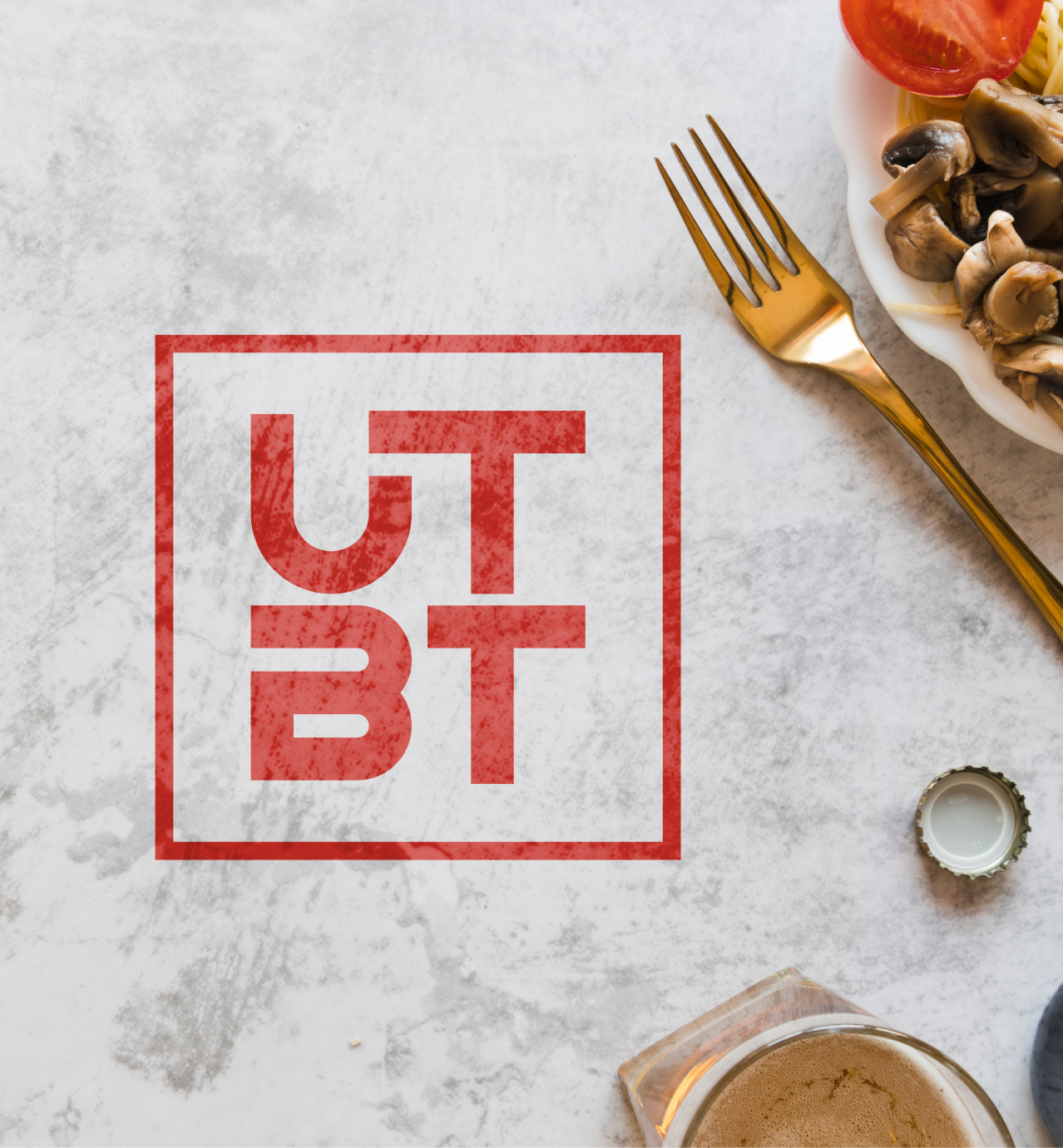 Red UTBT logo on a marble tabletop with a gold fork, plate of mushrooms and tomato, bottle cap, and a glass of beverage nearby.