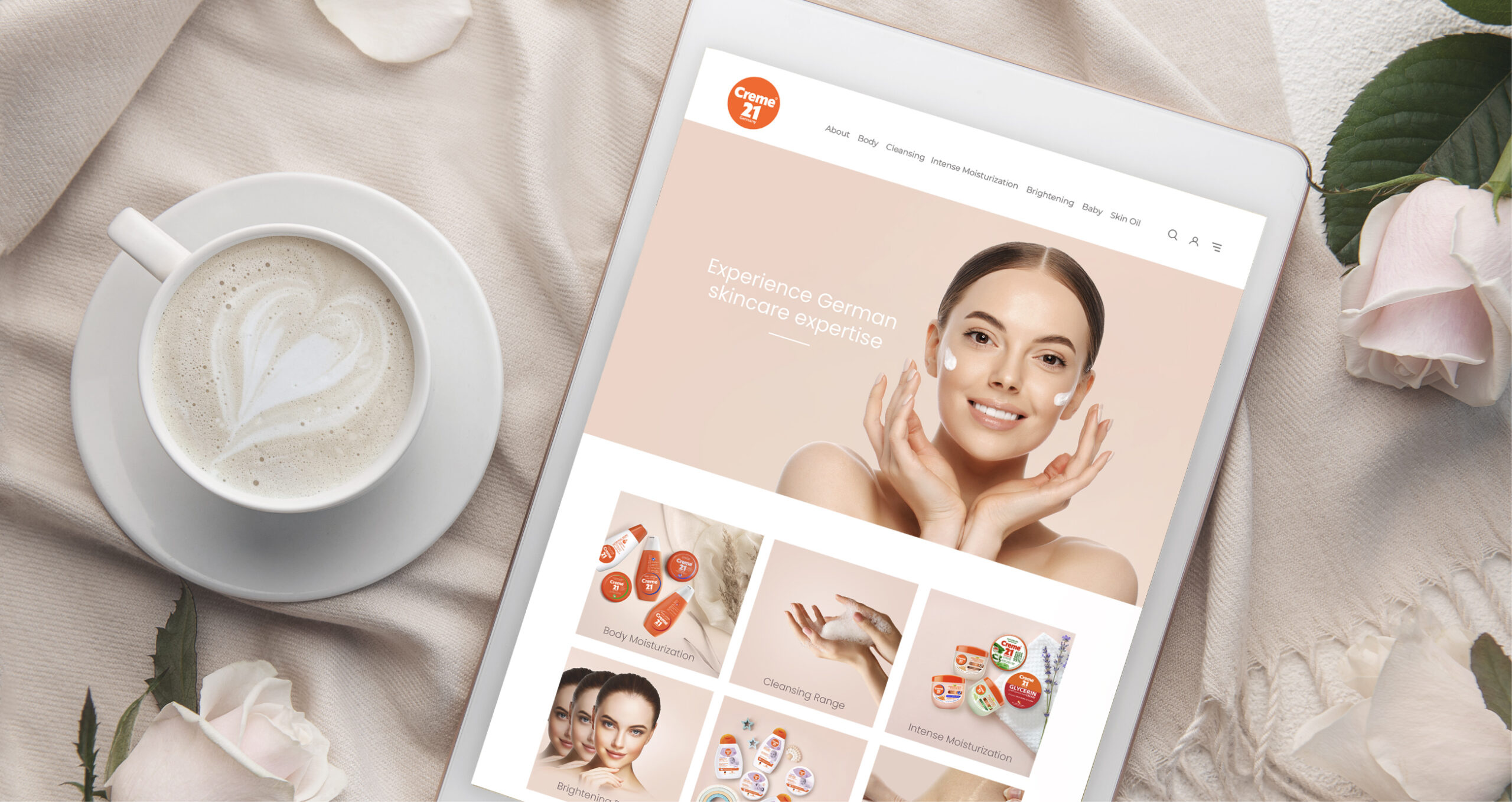 Tablet displaying Creme 21 skincare website with product categories, next to a cup of coffee and white roses on a soft fabric.