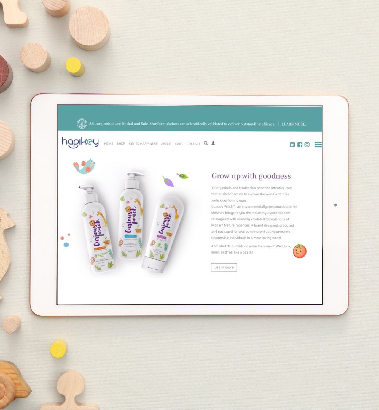 Tablet displays Hapikey website with kids' peach-themed skincare products and the slogan "Grow up with goodness," surrounded by toys.