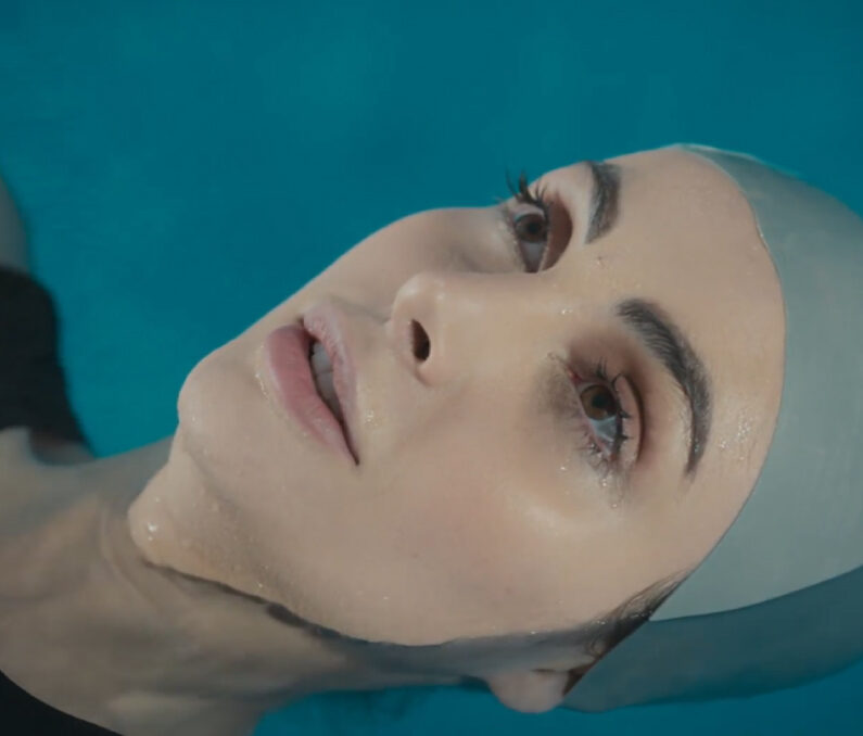 Woman floating in blue water with eyes open, peaceful expression, and droplets on her face.