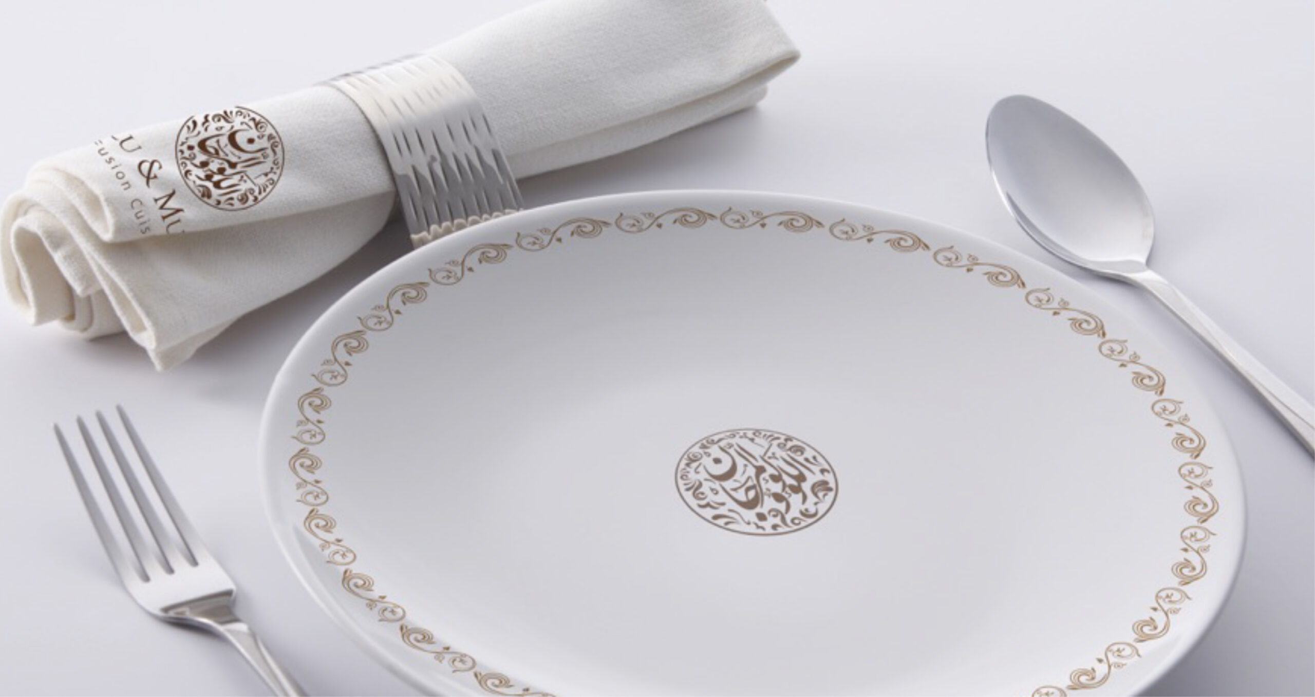 Elegant table setting with a white plate featuring gold Arabic calligraphy, a napkin with a silver ring, fork, and spoon.