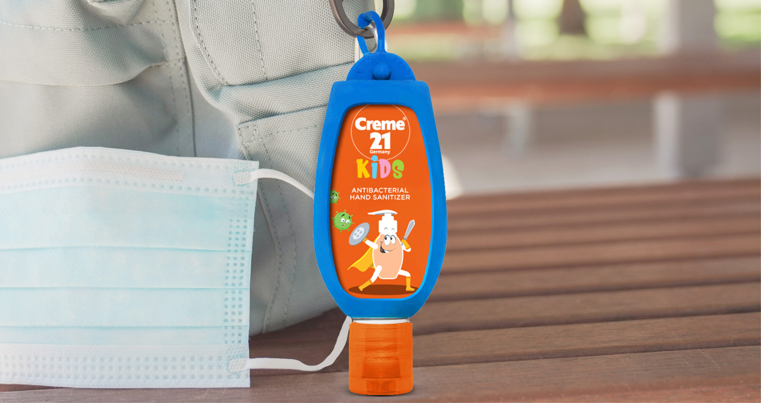 Creme 21 Kids antibacterial hand sanitizer in a blue holder clipped to a backpack, with a disposable face mask beside it.