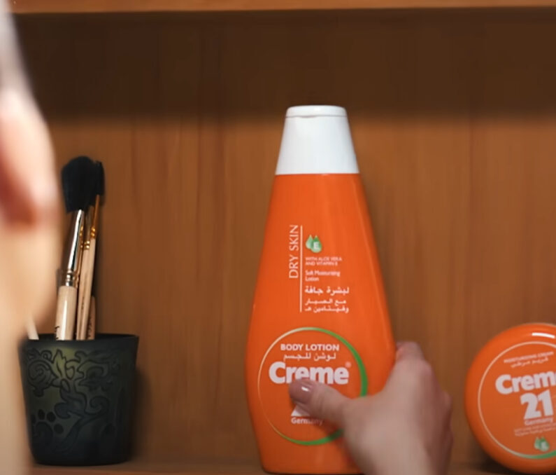 Creme 21 body lotion bottle being held with makeup brushes and skincare products on wooden surface.
