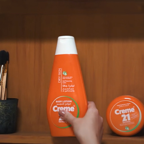 A hand picks up Creme 21 body lotion bottle from a shelf, next to a Creme 21 jar and brushes.
