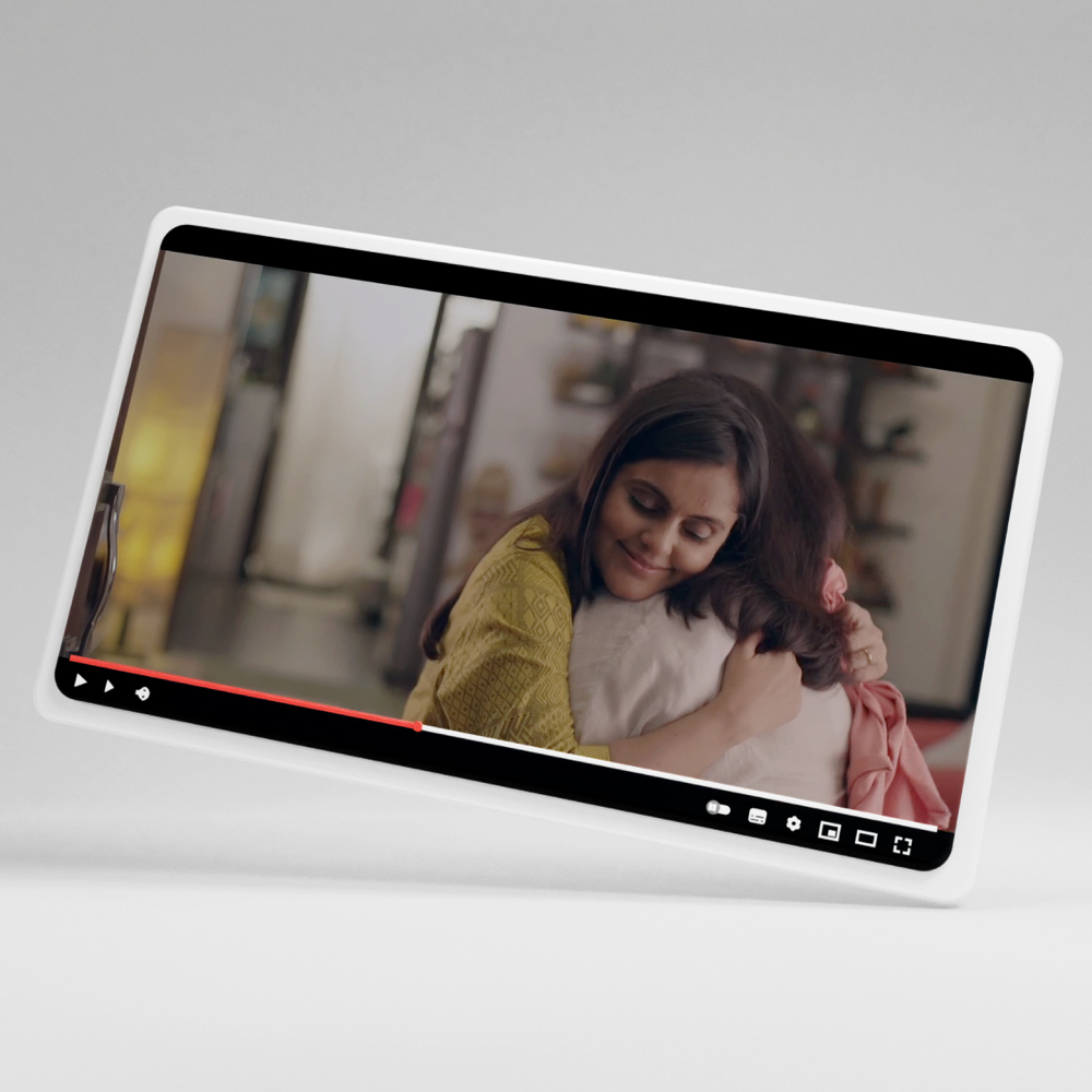 A woman in a yellow top hugs a white pillow closely while sitting in a cozy, softly lit home setting shown on a tablet screen.