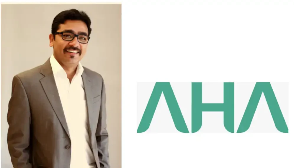 Man in a gray suit and white shirt standing next to a green AHA logo on a light background.