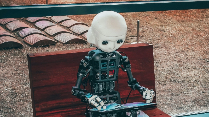 Humanoid robot with exposed mechanical parts sits on a bench using a tablet, near a window overlooking a grassy area.