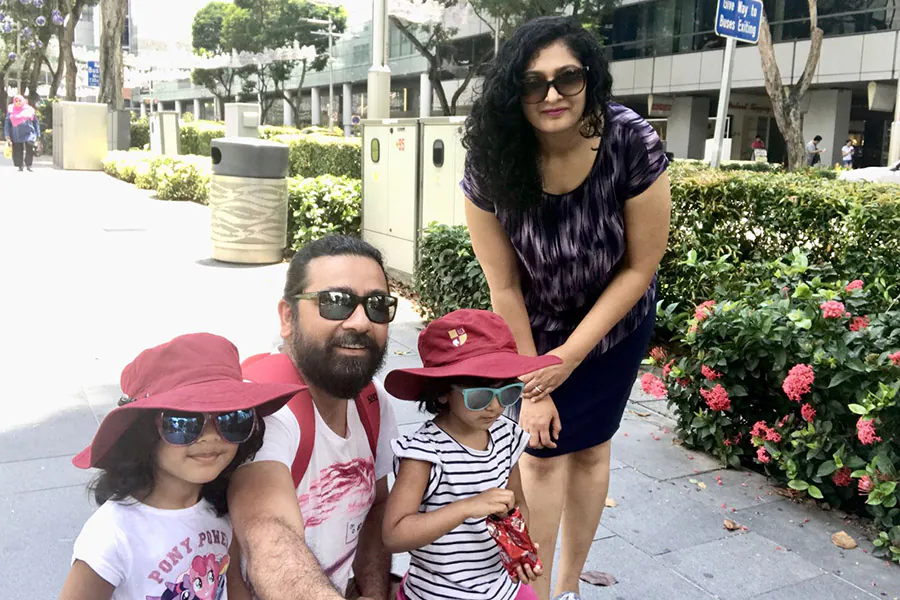Family with two children in red hats poses outdoors on a city sidewalk near bushes with red flowers and tall buildings.