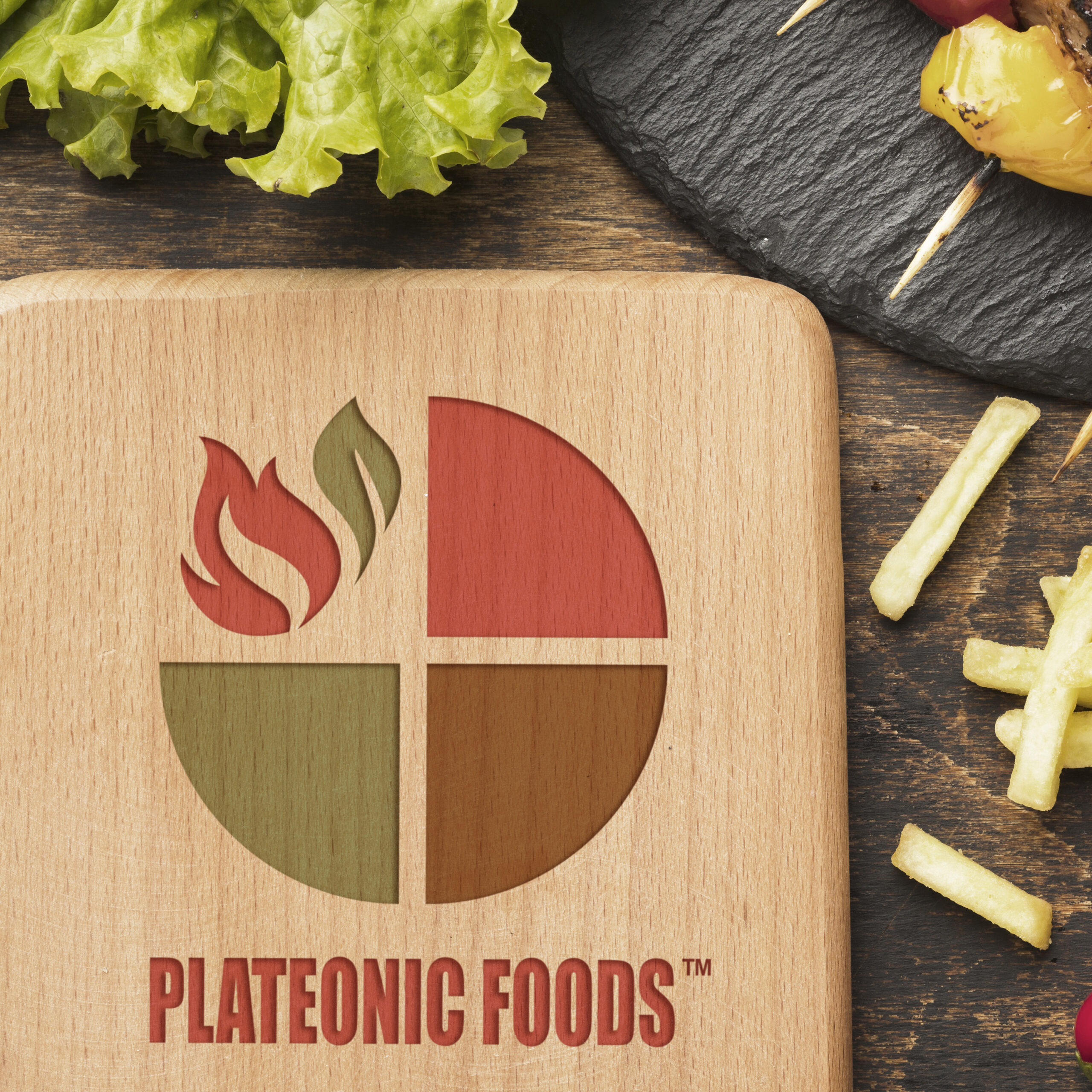 Plateonic Foods logo engraved on a wooden board with lettuce, fries, and grilled vegetables nearby.