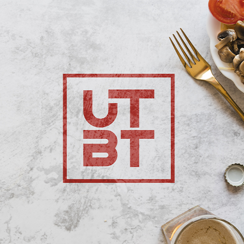 Red "UTBT" logo in a box on a marble table near a fork, plate with food, and a bottle cap.